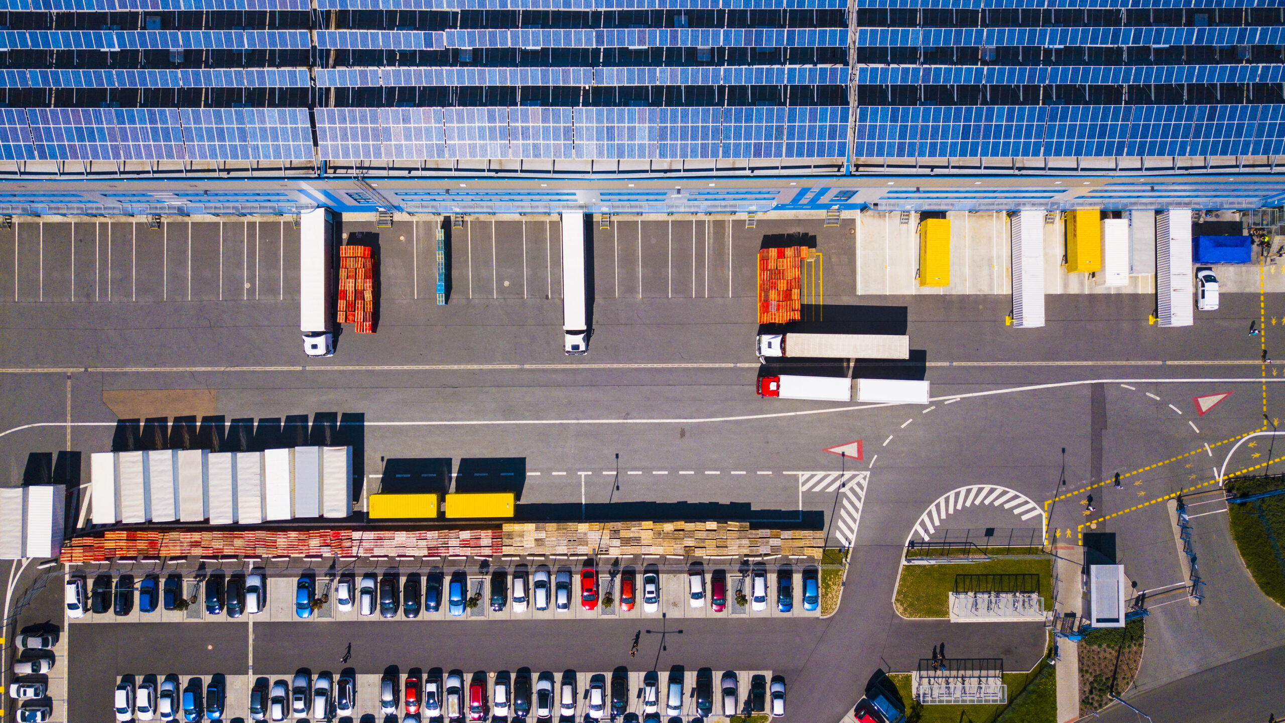 Aerial view of a solar panel covered roof of a distribution centre warehouse with adjacent truck loading bays, office block, and car parking area