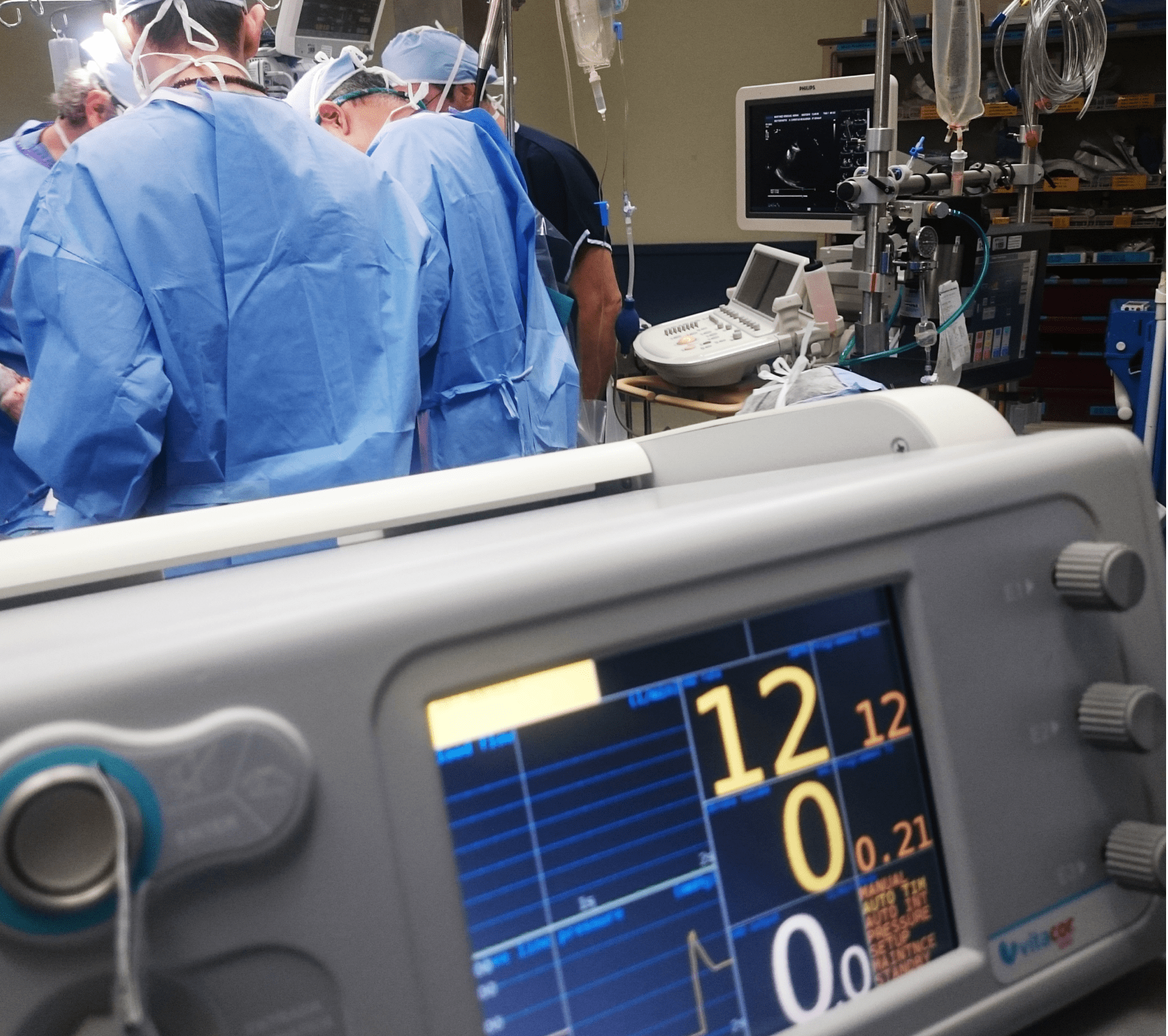 Close up view of a medical vital signs monitor in the foreground with a medical team working on a patient in a hospital theatre behind, their backs turned to the camera