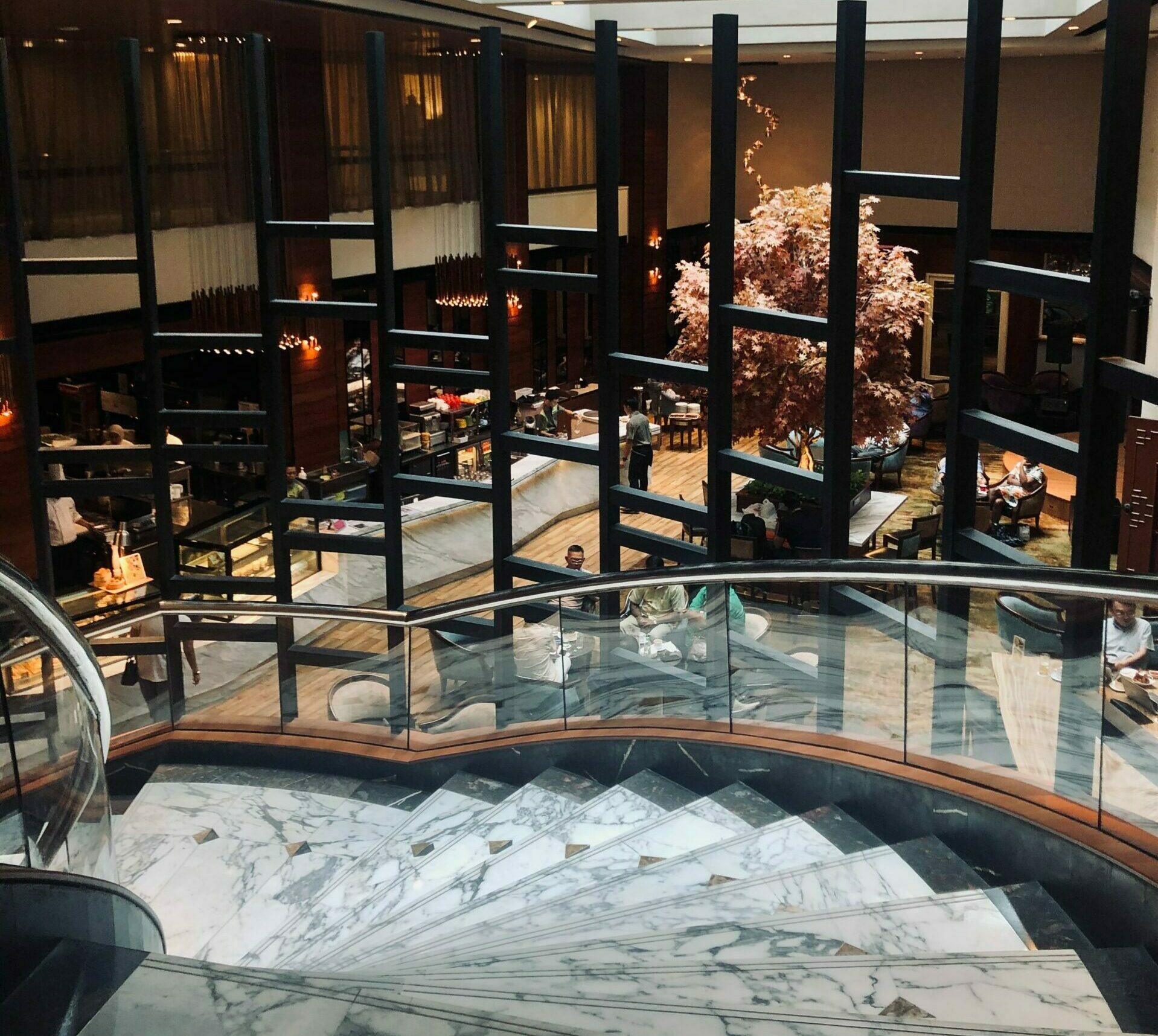 A curved staircase with stainless steel pipe handrails and glass partitioning looking down through a black square trellis at the reception area of a busy modern hotel